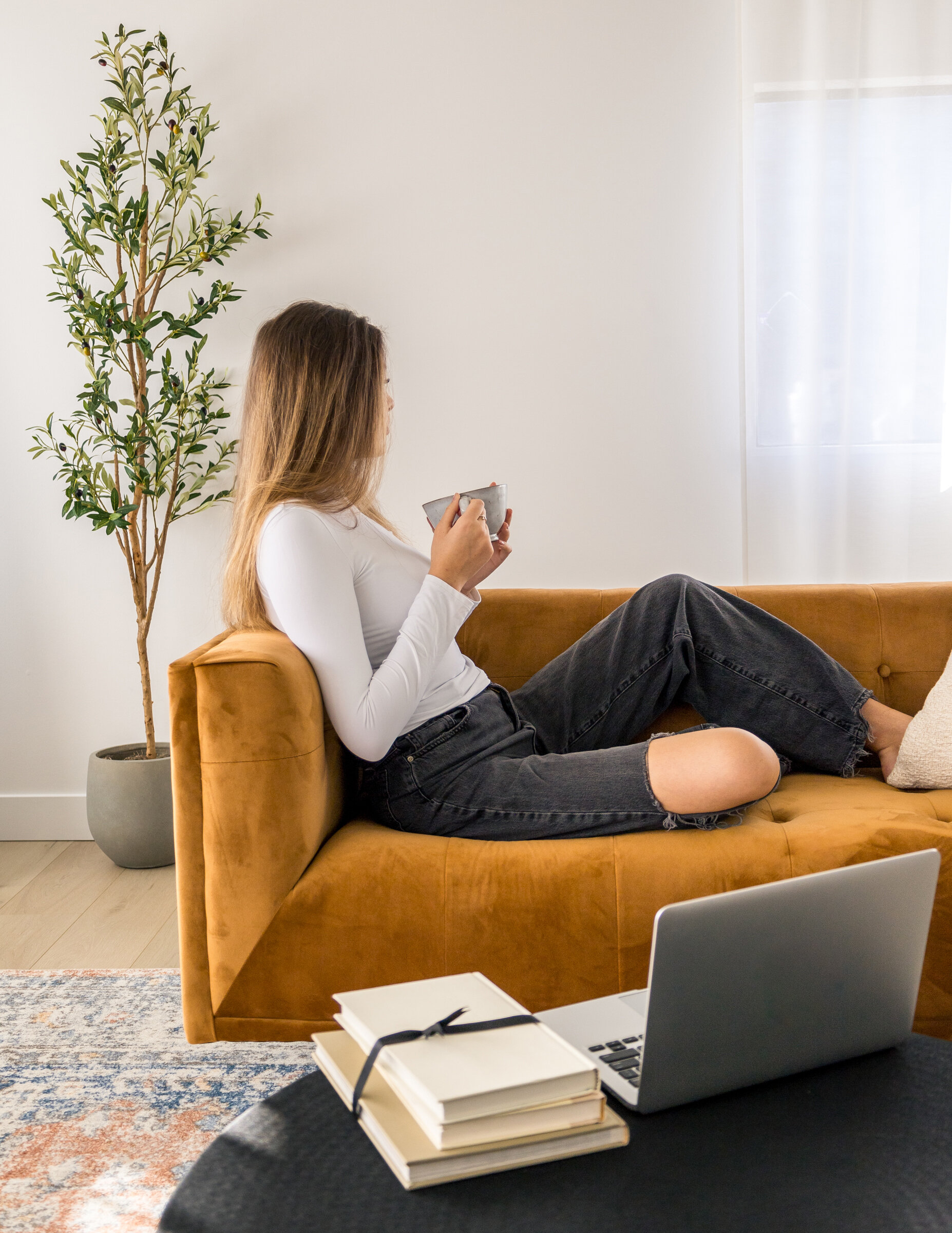 Live Authentically - woman with hole in knew pants sitting on orange couch drinking from a cup and staring out the window